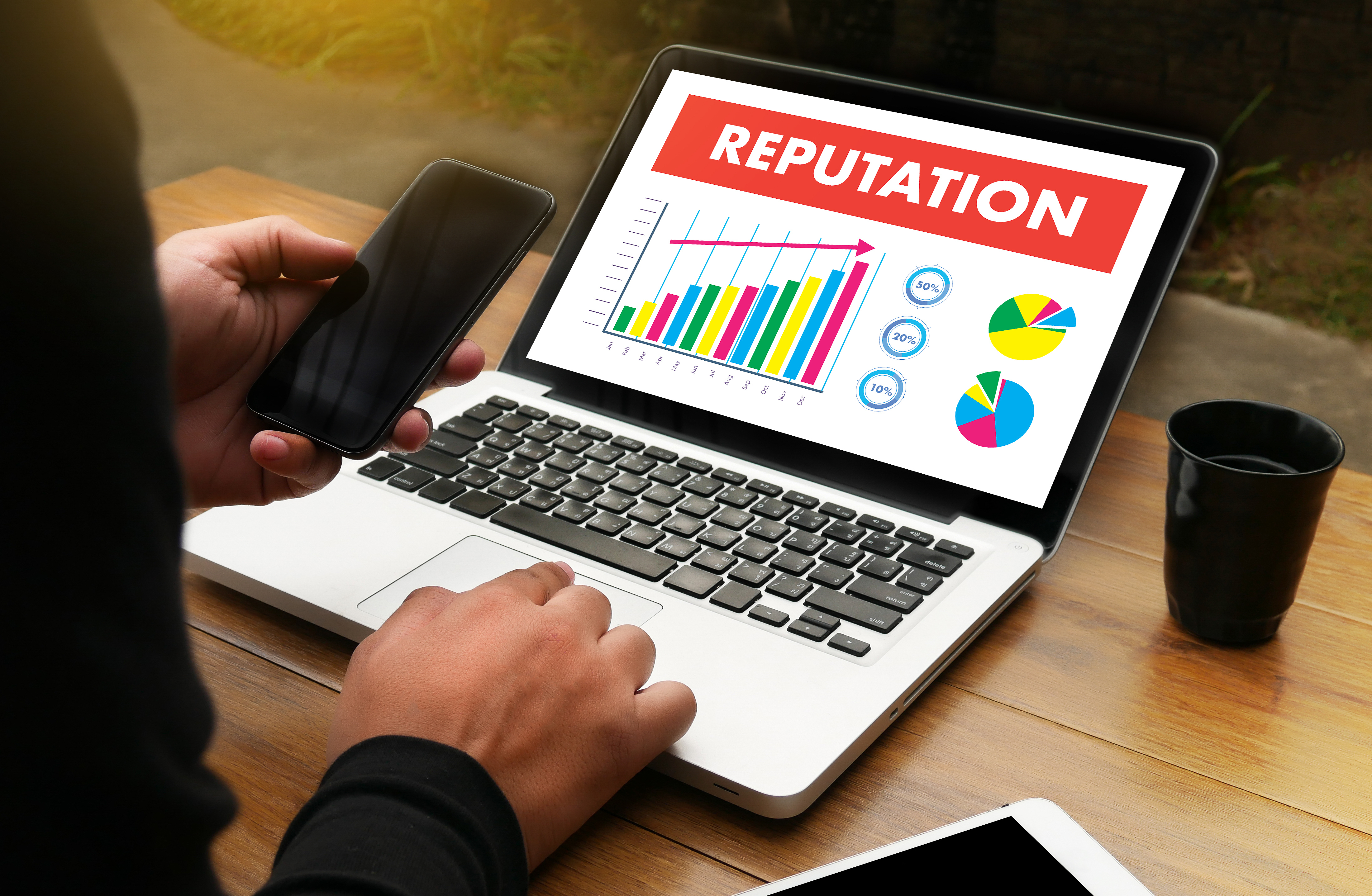 A person holding a phone on the left and typing on a laptop with right hand. The laptop has the word REPUTATION with graphs and charts below it.