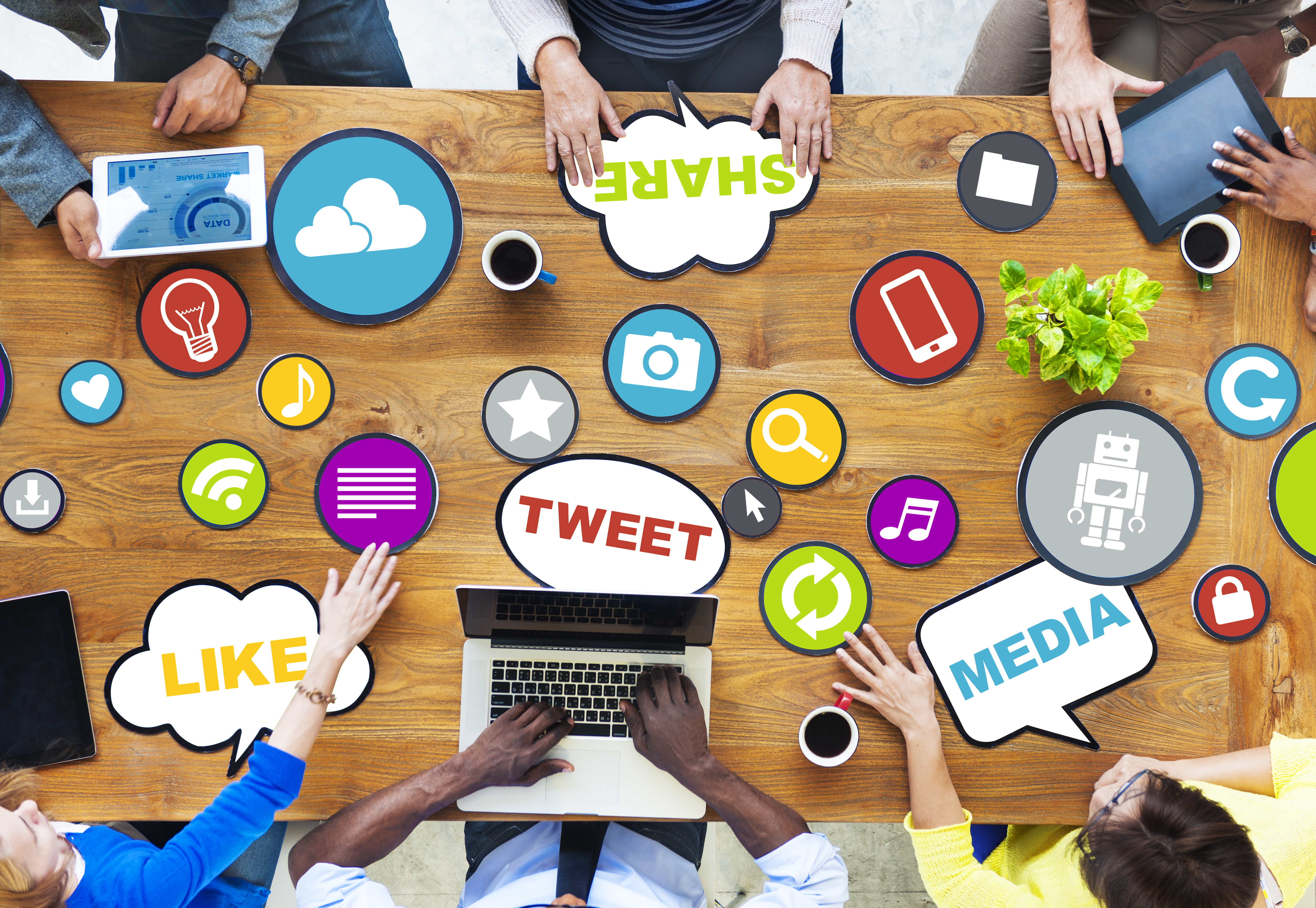 Table surrounded by people with laptops, ipads and printed social media icons of like, share, camera, media scattered on top